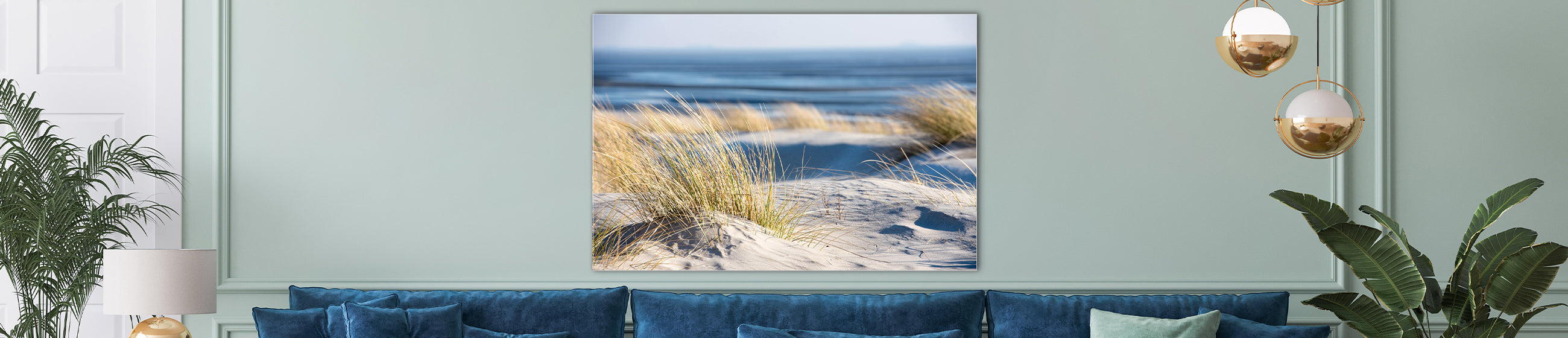 Fotokunst mit Dünen und Strandhafer im Vordergrund und Blick auf das Meer, aufgehängt in einem eleganten Wohnzimmer mit blauem Sofa und Pflanzen.